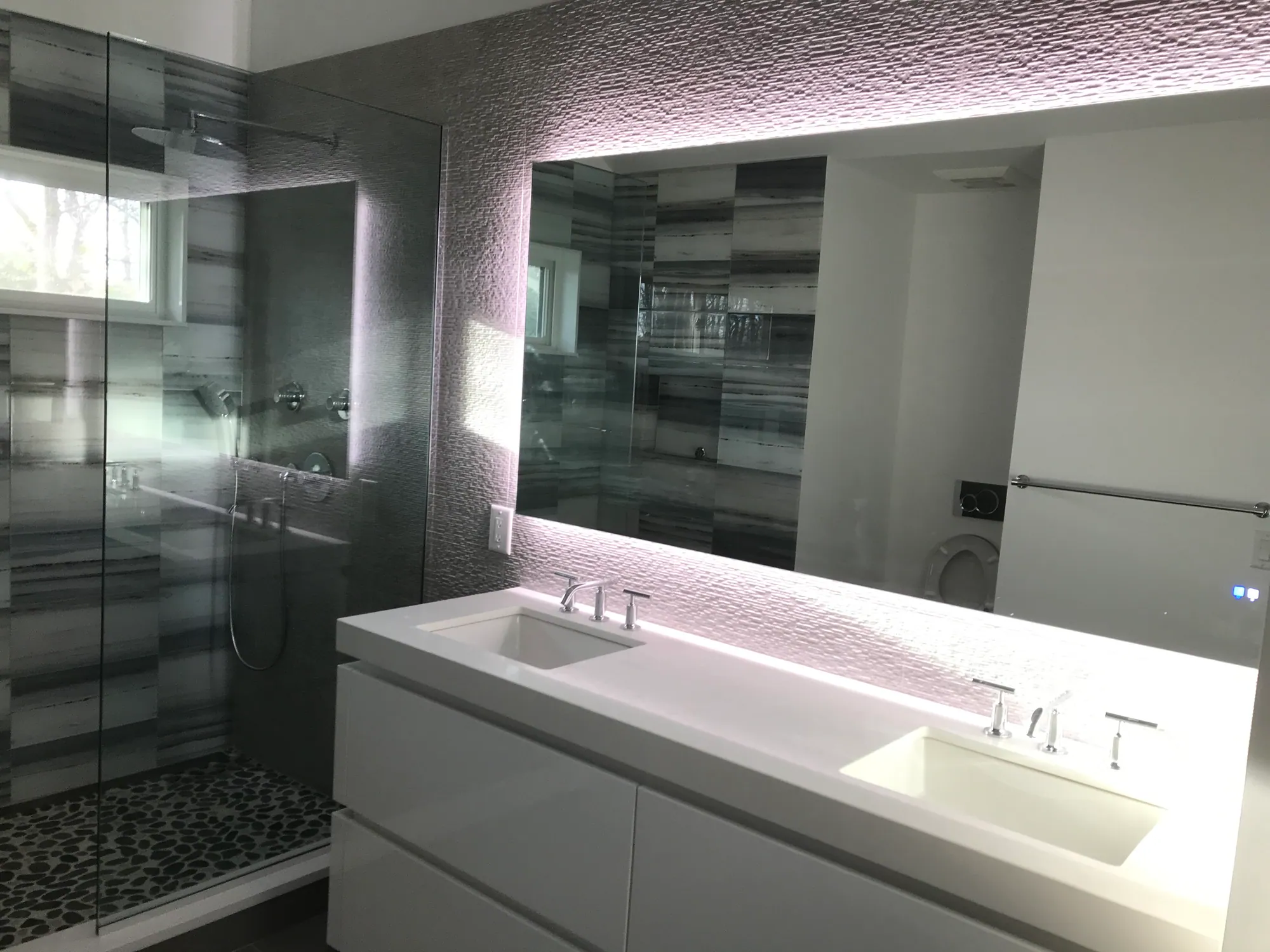 Renovated bathroom with a white floating double vanity, a large backlit mirror and a glass shower lined in grey striated stone-look tile.