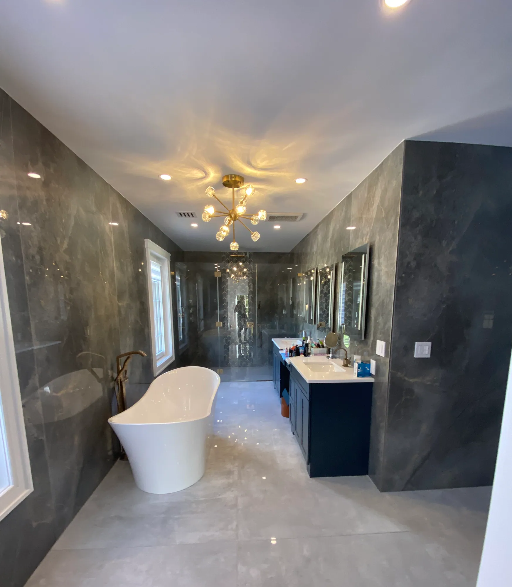 Large renovated primary bathroom with dark marble-look wall panels, a freestanding tub, a navy double vanity and a glass shower enclosure.