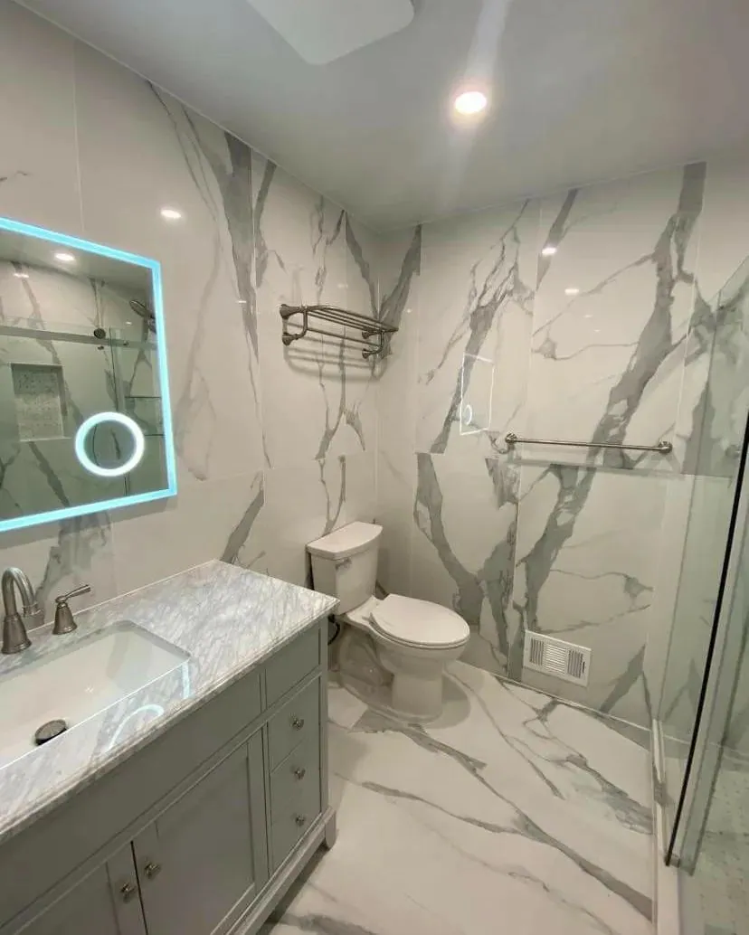 Renovated bathroom with large marble-look wall slabs, a backlit mirror, a grey vanity and a glass shower.