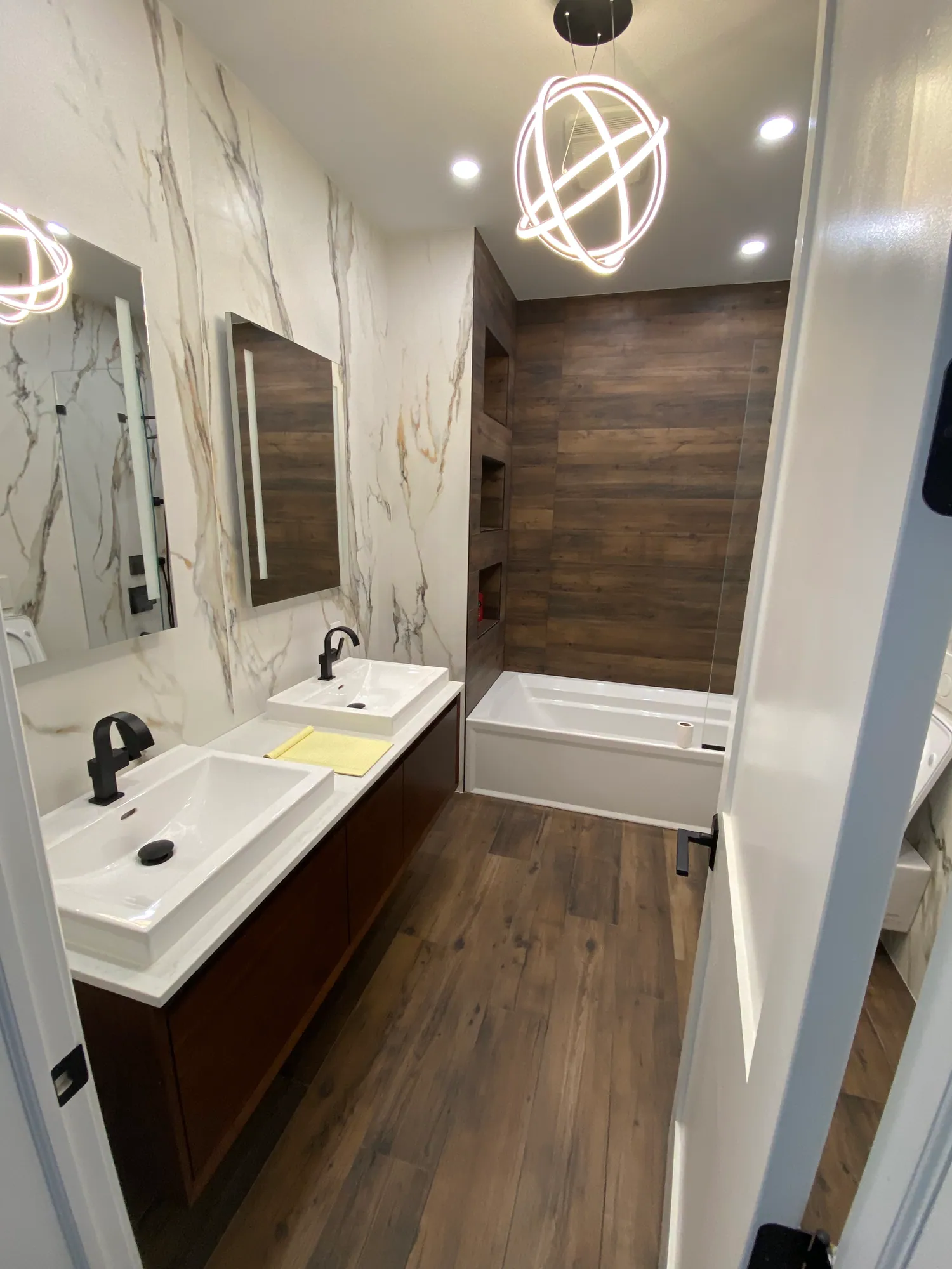 Renovated bathroom with a wood floating double vanity, marble-look wall tile, a wood-panel accent wall and a built-in tub.