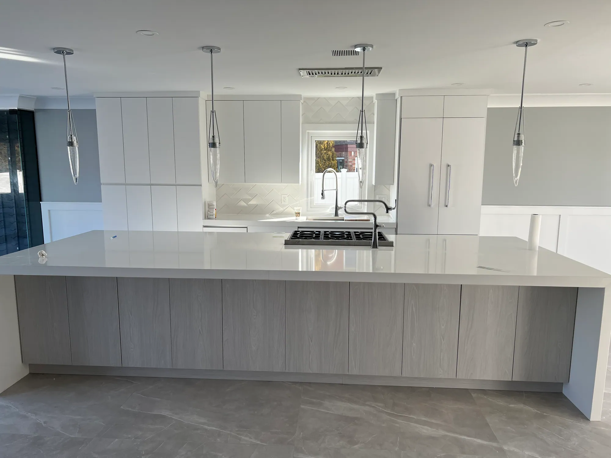 Modern kitchen renovation with handleless cabinets, a large island with a white countertop, herringbone tile backsplash and a built-in cooktop.