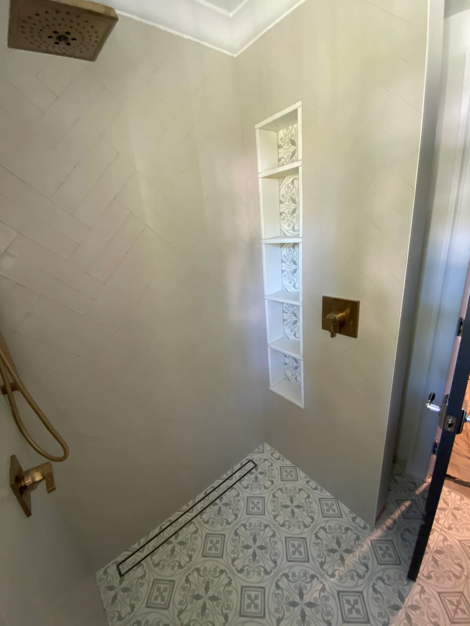 Walk-in shower with white wall tile, a tall decorative tile niche, brass fixtures and patterned floor tile.