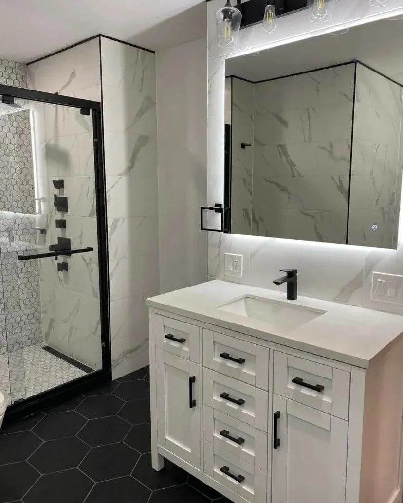 Walk-in shower with a black-framed glass enclosure beside a white vanity with a backlit mirror and black fixtures.
