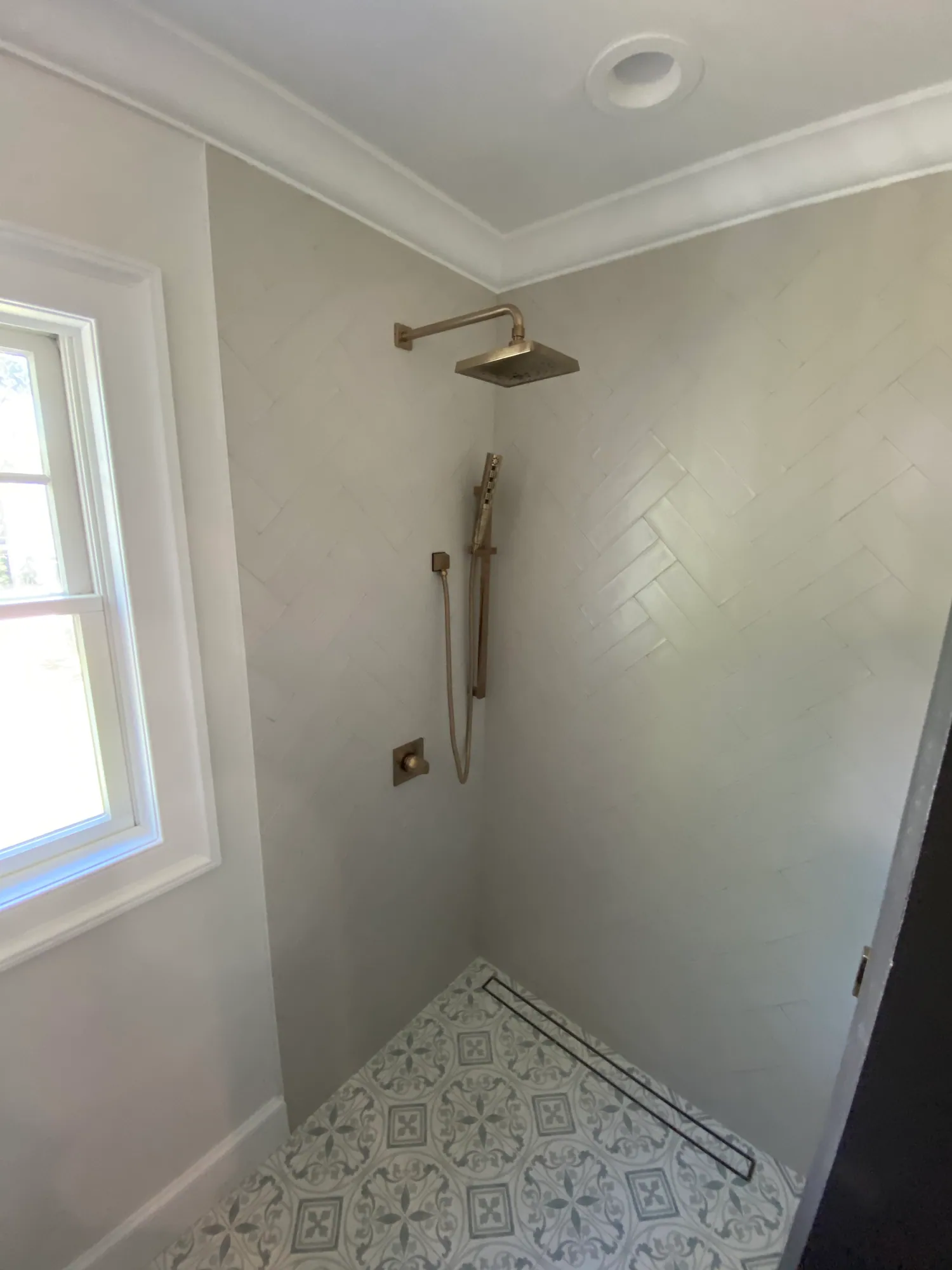 Walk-in shower with white wall tile, a brass rain head and hand shower, a window and patterned floor tile.